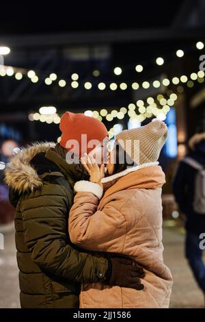 Vertikale mittlere Aufnahme von jungen asiatischen Mann und Frau in der Liebe tragen lässige warme Kleidung stehen im Freien am Winterabend küssen sich gegenseitig Stockfoto