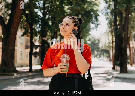 Ein Foto einer jungen Frau mit Zöpfen, die mit ihrem Telefon sprach, während sie einen Drink hielt Stockfoto