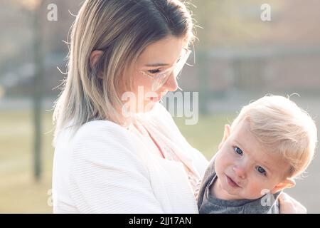 Glückliche junge Mutter mit einem kleinen Kind oder Kleinkind in den Armen, umarmend und kuschelend, stehend inmitten eines Parks in den Strahlen der aufgehenden Morgensonne. Hochwertige Fotos Stockfoto