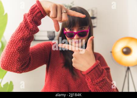 Kaukasische Frau mit dunklem Haar trägt eine rosa Sonnenbrille und einen roten Pullover, der das Gesicht mit den Händen umrahmt. Innenaufnahme. Hochwertige Fotos Stockfoto