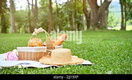 Sommer Picknick-Mittagessen im schönen grünen Garten, Picknickkorb mit Früchten und Gebäck, Strohpicknickhut, Blumenstrauß auf einer Picknick-Tischdecke. Stockfoto