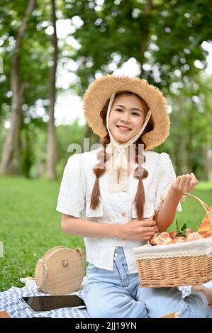 Portrait, Eine schöne junge asiatische Frau mit einem schönen Strohhut, sitzt auf einer Picknickdecke und hält einen Korbkorb im Garten. Stockfoto