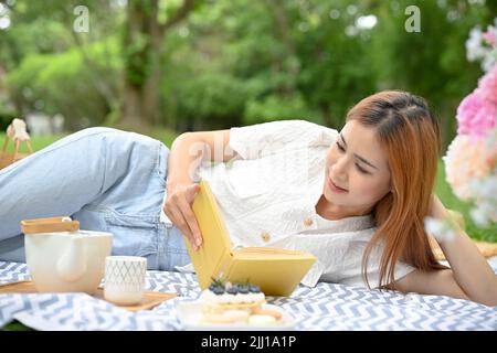 Eine hübsche junge asiatische Frau, die im Garten pickelt, sich auf ihrer Picknickdecke entspannt und dabei ihr Lieblingsbuch liest. Stockfoto