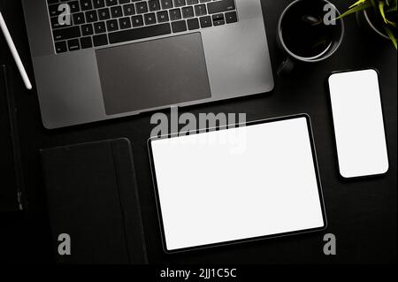 Moderner schwarzer Büroarbeitsplatz mit geöffnetem Notebook-Laptop, Kaffeetasse, Smartphone und digitalem Tablet-Modell mit weißem Bildschirm. Draufsicht, flach liegend Stockfoto