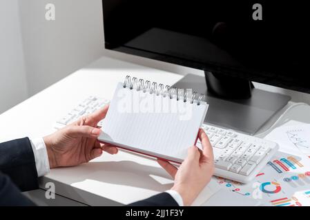 Geschäftsfrau Hält Notebook Mit Neuen Ideen Über Schreibtisch Mit Computer Und Tastatur. Frau Im Anzug Mit Notizpad Mit Wichtigen Nachrichten Über Office Stockfoto