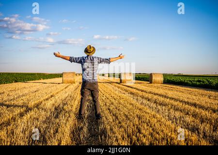 Glücklicher Bauer mit ausgestreckten Armen, der nach erfolgreicher Ernte neben Heuballen steht. Stockfoto