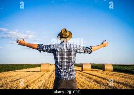 Glücklicher Bauer mit ausgestreckten Armen, der nach erfolgreicher Ernte neben Heuballen steht. Stockfoto