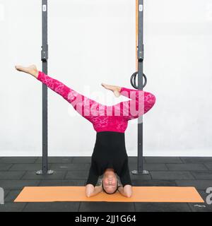 Blonde junge Frau Durchführung Kopfstand Yoga Pose mit den Füßen auseinander, aber eines der Beine gebogen. Lady in rosa Yoga-Leggings und schwarzem Shirt in einem Fitnessstudio. Stockfoto