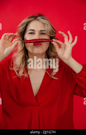 Vertikale lustige, kreative blonde Frau in rotem Outfit hält roten Paprika in den Händen unter der Nase wie Schnurrbart. Aktuelle Nachrichten Stockfoto
