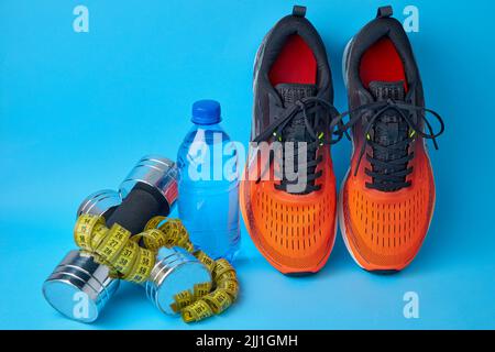 Orangefarbene Turnschuhe, Hanteln, Maßband und Trinkflasche aus Kunststoff auf blauem Hintergrund Stockfoto