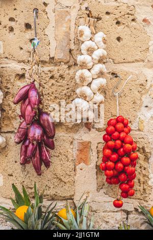 Bund und frischer weißer Knoblauch, rote Zwiebeln und Kirschtomaten hängen an einer Steinmauer in einem Straßenmarkt, vertikal Stockfoto