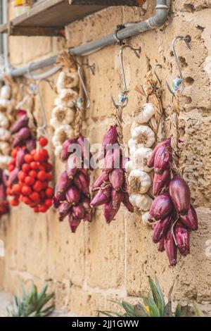 Bund und frischer weißer Knoblauch, rote Zwiebeln und Kirschtomaten hängen an einer Steinmauer in einem Straßenmarkt, vertikal Stockfoto