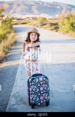 Die junge kaukasische Frau in einem Hut und Shorts steht mit einem roten Koffer auf der Straße. Stockfoto