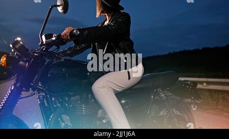 Hipster stilvolle Motorradfahrerin Frau sitzt auf Vintage-Stil Motorrad. Junge Fahrerin bei Nacht auf der Straße. Reise, Freiheit, klassisches Motorrad Stockfoto