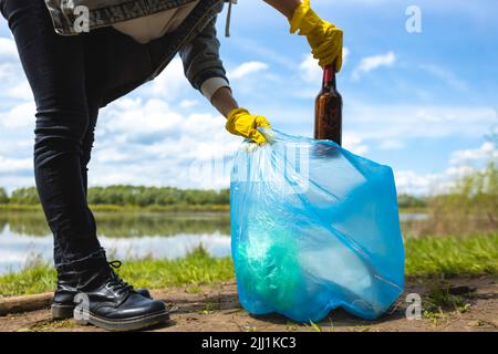 Eine junge Frau in gelben Handschuhen sammelt verlassenen Müll in einer schwarzen Tasche im Wald. Verschmutzung durch Plastik und Umweltschutz Stockfoto