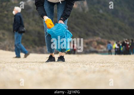 Eine Frau in Handschuhen mit einem speziellen blauen Beutel sammelt Müll im Sand entlang der Küste. Das Problem der Umweltverschmutzung. Müllabfuhr Stockfoto