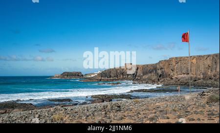 Eine rote Flagge flattert im Wind an der felsigen Küste des Ozeans gegen einen blau-bewölkten Himmel auf der Insel Teneriffa, Spanien Stockfoto