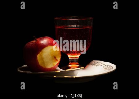 Roter Apfel mit einem Stück gebissen mit Glas mit roter Flüssigkeit auf zerbrochenem Porzellanteller Stockfoto