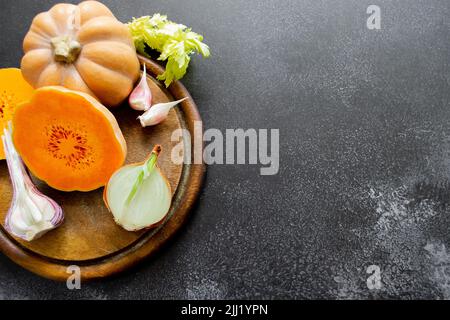 Frisches, helles Gemüse wird auf einem hölzernen Schneidebrett angelegt: butternusskürbis, grüne Sellerie-Blätter. Schwarzer ungleichmäßiger Hintergrund, Kopierbereich. Stockfoto
