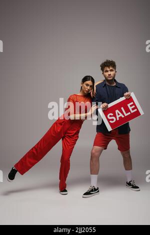 In voller Länge ein junges Paar, das Papier hält, mit einem Verkaufsbuchstaben auf Grau Stockfoto