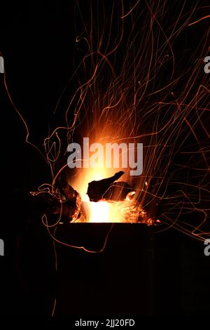 Feuerflammen und Funken aus Feuerholz, die nachts in einem Metallkamin vor dem dunklen Himmel brennen Stockfoto