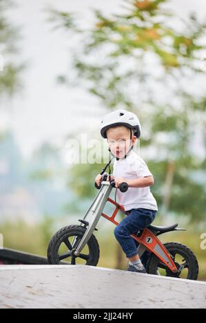 Porträt des Kindes Reiten Laufrad. Männliches Kleinkind im Helm, das im Skatepark lernt, auf dem Laufrad zu fahren. Stockfoto