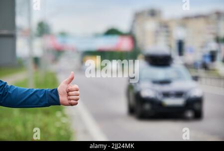 Nahaufnahme der Menschenhand, die am Straßenrand anhält. Männliche Hand zeigt Daumen nach oben Geste im Freien auf unscharfem Hintergrund. Anhalter, Anhalter, Auto-Stopp-Konzept. Stockfoto