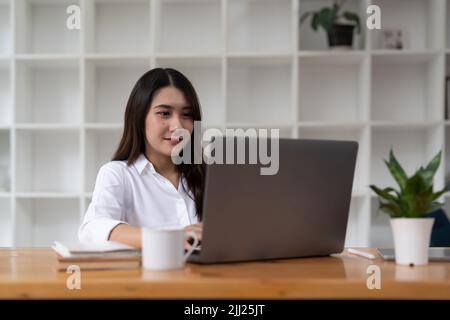 Junge glückliche asiatische Geschäftsfrau, die von zu Hause aus arbeitet und einen Computer-Laptop und eine Denkidee für ihr Geschäft nutzt Stockfoto