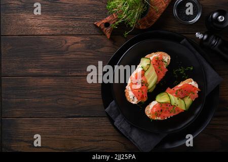 Sandwiches. Lachs-Toast mit Frischkäse, Gurke, schwarzem Sesam und Microgreens auf altem Holztisch-Hintergrund. Meeresfrüchte. Gesunde Ernährung. Fotografie Stockfoto
