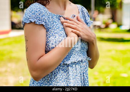 Süße Millennial-Frau in einem blauen Sommerkleid mit Blumenmuster, die im Stadtpark steht und im Freien gefaltete Hände auf der Brust hält. Dankbar sein und g zeigen Stockfoto