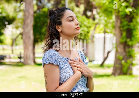 Nette Brünette Frau trägt Sommerkleid auf Stadtpark stehen, im Freien mit gefalteten Händen auf der Brust. Dankbar fühlen und Dankbarkeit zeigen Zeichen, b Stockfoto