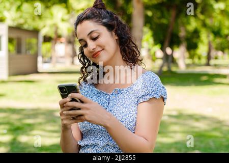 Junge Frau, die selbstbewusst lächelt und Sommerkleid trägt, steht im Stadtpark, schaut im Freien auf den Telefonbildschirm und benutzt das Telefon. Nachrichten an Freunde senden Stockfoto