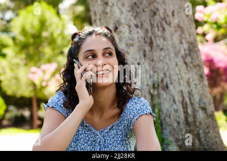 Junge Frau, die selbstbewusst lächelt, trägt Sommerkleid und steht im Stadtpark, spricht im Freien mit Freunden oder ihrem Freund mit einem Lächeln auf dem Handy. Stockfoto