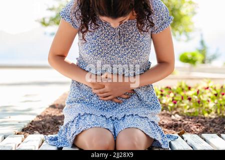 Junge schöne Frau trägt Sommerkleid im Stadtpark, im Freien mit der Hand auf dem Bauch, weil Verdauungsstörungen, schmerzhafte Krankheit fühlen sich unwohl. Schmerzen oder Pr Stockfoto