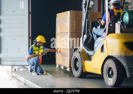 Asiatischer Gabelstaplerfahrer, der einen Frachtcontainer mit einer vollen Palette mit Boxen im Logistikhafen verlädt. Asiatischer Lagerarbeiter und Sicherheit i Stockfoto