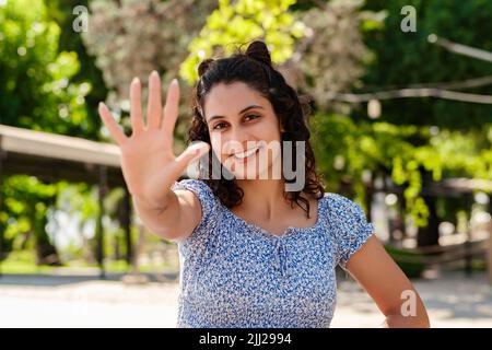 Junge Frau, die selbstbewusst lächelt und Sommerkleid trägt, steht im Stadtpark, zeigt sich im Freien und zeigt mit den Fingern Nummer fünf auf, während sie conf lächelt Stockfoto