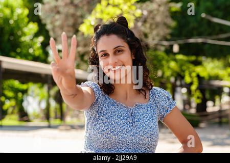 Fröhliche Brünette Frau trägt Sommerkleid im Stadtpark, zeigt im Freien und zeigt mit den Fingern Nummer drei, während sie selbstbewusst lächelt und happ Stockfoto