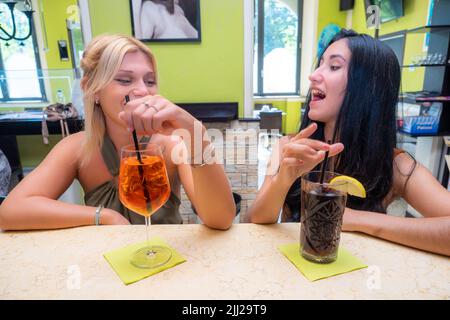 kaukasische blonde Frau trinkt Spritz in der Nähe einer Latino-Frau, die in der Kneipe Cola trinkt. Stockfoto