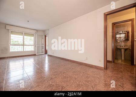 Leeres Wohnzimmer mit rötlichem Steinzeugboden, weiß gestrichenen Wänden, Fenstern und Ausgang zu einer Terrasse einer Wohnung Stockfoto