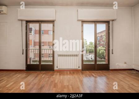 Leeres Wohnzimmer mit Eichenholzboden, weiß gestrichenen Wänden und zwei Fenstern, die zu einem länglichen Balkon mit Aussicht führen Stockfoto