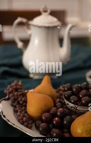 Auf einer dunkelgrünen Tischdecke vor einer teekannen aus china sitzt eine weiß-silberne Platte aus violetten Muscadinen, winzigen Trauben und Bosc-Birnen. Stockfoto