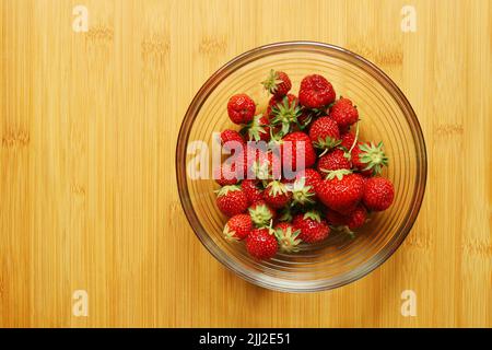 Frische, saftige Bio-Erdbeeren mit Blättern in der runden Glasschale, die auf das Holz-Bambus-Schneidebrett gelegt wird. Zutaten für Marmelade, Kuchen, Dess Stockfoto