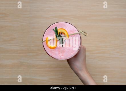 Frau hält ein Glas gemischten Beerenobst-Smoothie und Orangenschale auf einem Holztisch in der Hand Stockfoto