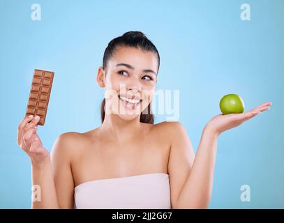 Shes hat eine Entscheidung getroffen. Studio schoss eine attraktive junge Frau, die sich vor blauem Hintergrund zwischen einer Schokolade und einem Apfel entschied. Stockfoto