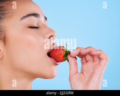 Shes in Liebe mit Gesundheit. Studioaufnahme einer attraktiven jungen Frau, die vor blauem Hintergrund in eine Erdbeere beißt. Stockfoto