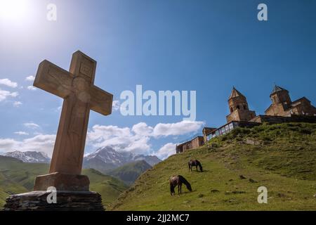 Steinernes orthodoxes Kreuz mit einer Gergeti-Trinity-Kirche mit weidenden Pferden auf dem grünen Grashügel und dem Berg Kazbek 5054m. Weitwinkellandschaft nahe Th Stockfoto