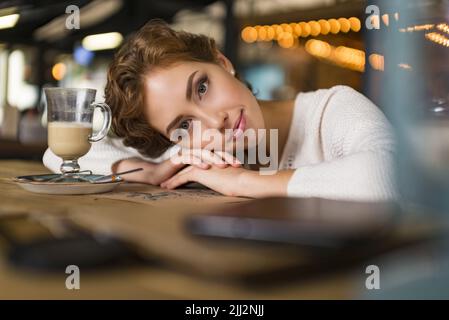 Junge nachdenkliche Frau in einem Café. Sie legte ihren Kopf in die Hände und schaute ruhig und friedlich auf die Kamera. Das Konzept der Psychologie und der inneren Harmonie. Stockfoto