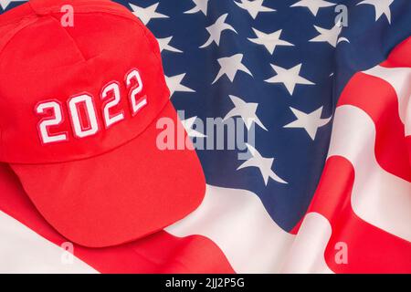 Rote MAGA-Kappe mit Datum 2022 und US Stars and Stripes-Flagge. Für die US-Zwischenwahlen vom 2022. November und die Ausrottung der Demokraten durch die US-Republikaner der Roten Welle. Stockfoto