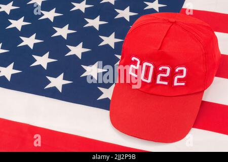 Rote MAGA-Kappe mit Datum 2022 und US Stars and Stripes-Flagge. Für die US-Zwischenwahlen vom 2022. November und die Ausrottung der Demokraten durch die US-Republikaner der Roten Welle. Stockfoto