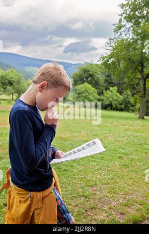 Kleiner Junge, der eine Karte, ein Papier, Anweisungen in einem Berghochland, Wald hinter sich liest. Auf einer Wiese, Besuch, Urlaubsreise, tourisme. Zehn Jahre alt Stockfoto